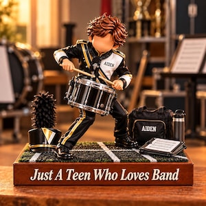 Op de afbeelding: Een beeldje van een drummer in een zwart-wit uniform van een marchingband met gouden accenten, spelend op een kleine trommel. De basis bevat de tekst 'Just A Teen Who Loves Band', een zwarte hoed, een tas met 'AIDEN' en een waterfles.