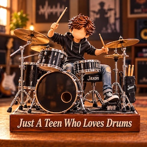 Op de afbeelding: Een beeldje van een persoon met bruin haar in een zwarte hoodie en blauwe spijkerbroek die op een zwart drumstel speelt. De drumkruk en een zijtas zijn gelabeld met Jaxon. De houten voet bevat de tekst Just A Teen Who Loves Drums in het wit.