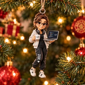 May include: A hanging ornament featuring a brown-haired character with glasses, wearing a light gray hoodie, black pants with the name ZOE and a heart printed on the leg, and white sneakers. The character holds a laptop and a mug with code symbols.