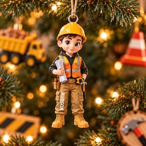 May include: A hanging ornament depicting a construction worker figure wearing a yellow hard hat, an orange reflective safety vest, and tan cargo pants with the name HUDSON printed on the leg. The figure holds a rolled blueprint and a hammer.
