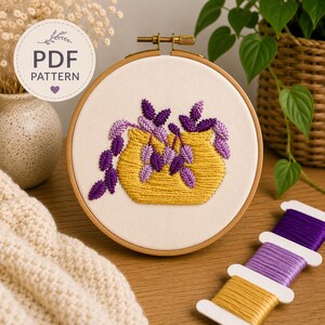 May include: Embroidered floral design in a wooden hoop. The artwork features a yellow pot with purple flowers. The text "PDF PATTERN" is in a circle. Spools of purple and yellow thread are visible.