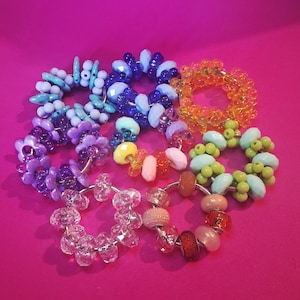 May include: A selection of colourful beaded bracelets in diverse designs. The bracelets showcase a variety of beads in shades of blue, purple, orange, green, pink, and clear. The beads are strung on a silver wire, creating a vibrant and eye-catching accessory.