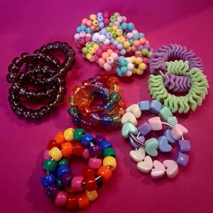 May include: A selection of beaded bracelets and rings in various colours. The bracelets feature diverse designs, including rainbow-coloured beads, heart-shaped beads, and spiral rings. The colour palette includes shades of pink, purple, green, orange, and blue.