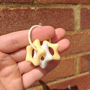May include: A set of star-shaped teething rings in shades of yellow and white, attached to a cream-coloured ring. The rings are held against a brick wall background.