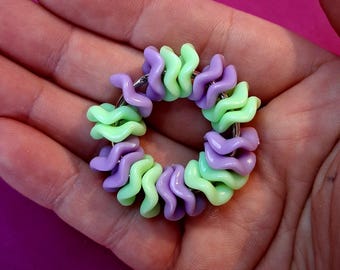May include: A small, circular keychain featuring a spiral pasta design. The keychain is composed of alternating light green and purple spiral beads. The beads are strung together on a silver-colored metal ring. The keychain is held in a person's hand.