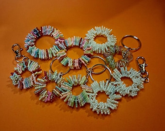 May include: A collection of beaded keychains in various colors, including white, pink, blue, and green. The keychains are circular and feature a variety of bead sizes and textures. Some have silver key rings and lobster claw clasps.