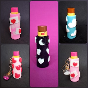 May include: A selection of decorative lip balm tubes in different colours. The tubes feature designs such as hearts, clouds, and celestial patterns. Some tubes have keychains with glitter-filled charms. The caps are pink or brown.