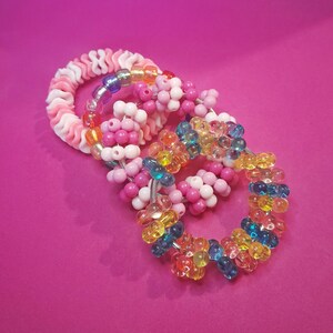 May include: Three colourful beaded bracelets on a pink background. One bracelet is white and pink with a wavy design. The other two are circular, with pink, white, and rainbow-coloured beads. The third bracelet is made of clear, yellow, blue, and orange beads.