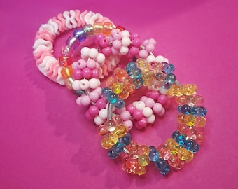 May include: Three colorful beaded bracelets on a pink background. One bracelet is white and pink with a wavy design. The other two are circular, with pink, white, and rainbow-colored beads. The third bracelet is made of clear, yellow, blue, and orange beads.