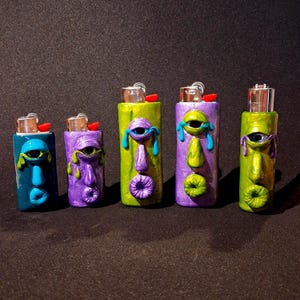 May include: Five decorative lighters with sculpted faces in various colours. The lighters are painted in shades of blue, purple, and green, with expressive facial features. Each lighter has a different face design, with eyes, noses, and mouths.