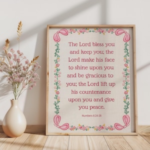 May include: Framed artwork with a light wood frame. The artwork features a cross-stitch design with the text "The Lord bless you and keep you..." in pink. The border is decorated with pink bows and floral accents. A vase with flowers is to the left.