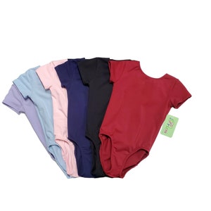 May include: Five short-sleeved leotards in different colors: light blue, pink, navy blue, black, and red. The leotards are arranged in a semi-circle, with the red leotard in the front. The leotards have a scoop neck and a fitted bodice. The leotards are made of a soft, stretchy fabric.