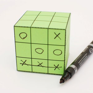 May include: A green paper cube with a tic-tac-toe game drawn on it. The cube is divided into nine squares, and the game is in progress. There is a black marker pen next to the cube.