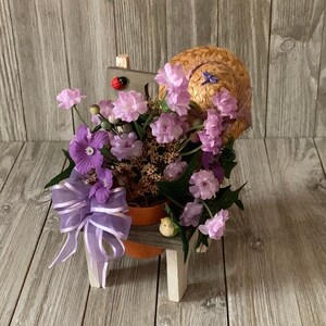 May include: A purple flower arrangement in a terracotta pot, sitting on a small wooden stool with a straw hat on top. The stool is painted white and has a small bird figurine on the side.