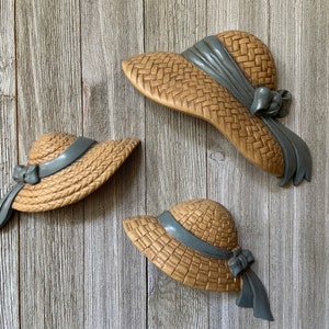 May include: Three straw hats with blue ribbons hanging on a wood wall. The hats are different sizes and are all in the same style.