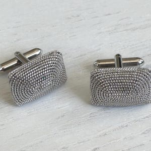 May include: A pair of rectangular silver-toned cufflinks featuring a textured, woven metal surface pattern. Each cufflink has a polished silver-toned swivel toggle mechanism on the back, designed to secure through the buttonholes of a shirt cuff.