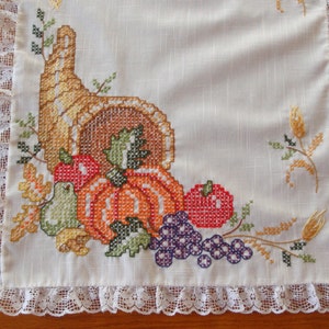 May include: Cream-colored, embroidered table runner with a harvest theme. Features a cornucopia overflowing with a pumpkin, apples, grapes, and wheat. The edges are trimmed with a delicate, white lace.