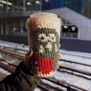 May include: A crocheted coffee cup cozy in cream, green, and red. The cozy features a design with a green logo and a red heart. The cup has a clear plastic lid and is held in front of a snowy outdoor scene.