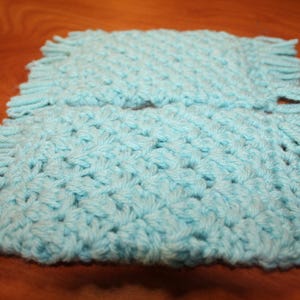 May include: Two light blue crocheted square mats with fringed edges. The mats are made of soft yarn and have a textured, woven appearance. The mats are displayed on a wooden surface.