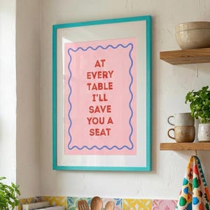 以下が含まれることがあります： 「AT EVERY TABLE I'LL SAVE YOU A SEAT」の赤い文字がピンク色の背景に描かれた額入りのプリント。ターコイズのフレームが特徴です。カラフルなタイル、木製の棚、フルーツボウルなど、活気があり、魅力的な雰囲気を作り出すキッチンシーンです。