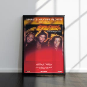 May include: A framed poster featuring the Bee Gees album Spirits Having Flown. The design includes a photograph of the three band members, the album title, and a tracklist with songs like Tragedy and Too Much Heaven, set against a red background.