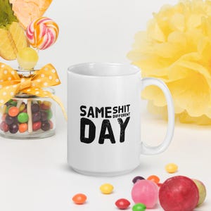 May include: White ceramic mug with the text "SAME SHIT DIFFERENT DAY" in black. The mug is surrounded by colourful sweets and a yellow paper flower.