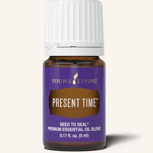 Puede incluir: Pequeña botella de vidrio marrón con tapa blanca, etiquetada "PRESENT TIME" en texto blanco sobre fondo morado. Contiene una mezcla de aceites esenciales premium, 5 ml. También se ve la marca "YOUNG LIVING".