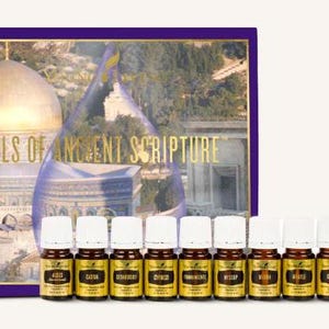May include: A collection of essential oil bottles with white lids and gold labels, displayed in front of a book. The book cover has a purple border and gold lettering that reads "Oils of Ancient Scripture", with a background image of a building.