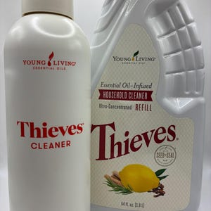 Könnte beinhalten: Zwei weiße Flaschen Thieves Cleaner von Young Living. Eine ist eine Sprühflasche, die andere eine Nachfüllflasche. Die Nachfüllflasche hat ein Etikett mit Zitrone, Zimtstangen und Nelken. Die Flaschen enthalten 1,8 Liter Reiniger.