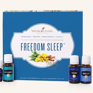 May include: A product box with the text "FREEDOM SLEEP" and the brand name "Young Living". The box is blue with a white label. There are four small essential oil bottles in front of the box. The bottles have white caps and blue labels.