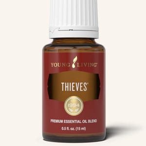 Op de afbeelding: Een bruine glazen fles van Young Living Thieves essentiële olie blend. De fles heeft een witte dop en een rood etiket met het woord "THIEVES" in goud. Het etiket bevat ook de tekst "PREMIUM ESSENTIAL OIL BLEND" en "15 ml".