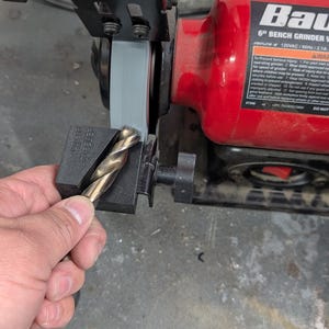 May include: A bench grinder with a red housing and a gray grinding wheel. A gold-colored drill bit is held in a black drill bit sharpener. The grinder has the text "Bau" and "6" BENCH GRINDER W" on it.