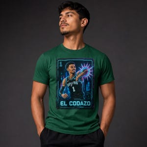 May include: Green t-shirt featuring a graphic of a basketball player in a black and white jersey with the number 1. The graphic is framed with a neon blue border and the text "EL CODAZO".
