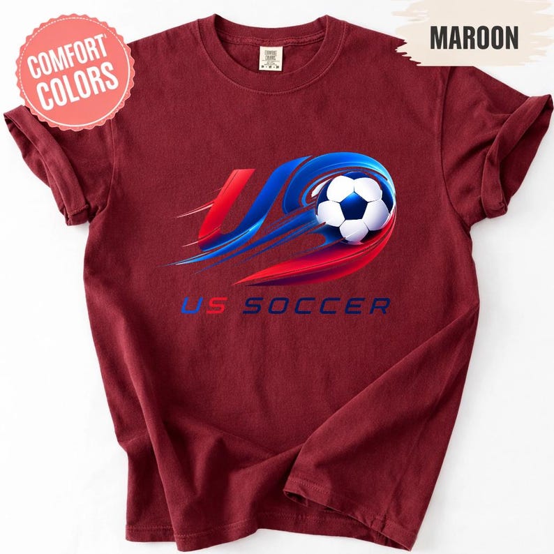 K&ouml;nnte beinhalten: Ein kastanienbraunes Kurzarm-T-Shirt mit einer Grafik eines Fu&szlig;balls, der von roten und blauen Bewegungsstreifen umgeben ist. Der Text US SOCCER ist in Blau unter der Grafik gedruckt. Ein Comfort Colors-Etikett ist am Halsausschnitt sichtbar.