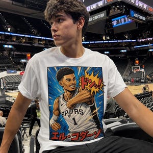 May include: White t-shirt featuring a graphic of a basketball player in a Spurs jersey with the number 1. The graphic has a comic book style with Japanese text. The shirt is worn by a person in a stadium setting.