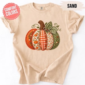 Könnte beinhalten: Ein sandfarbenes T-Shirt mit einer großen, zentralen Grafik eines Kürbisses, der aus verschiedenen gemusterten Stoffsegmenten besteht, darunter orangefarbenes Vichy-Karo, Blumenmuster und grüne Punkte, mit einem braunen Stiel und grünen Blättern.