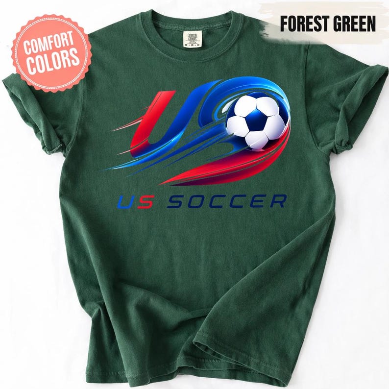 K&ouml;nnte beinhalten: Ein waldgr&uuml;nes T-Shirt mit einer Grafik eines schwarz-wei&szlig;en Fu&szlig;balls mit roten und blauen Bewegungsstreifen. Der Text US SOCCER ist unter der Grafik in Blau und Rot gedruckt. Ein Comfort Colors-Etikett ist am Halsausschnitt sichtbar.