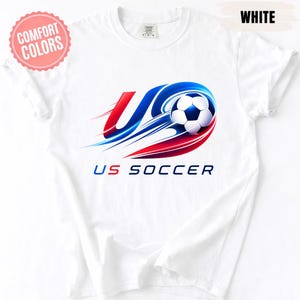 K&ouml;nnte beinhalten: Ein wei&szlig;es Kurzarm-T-Shirt mit einer Grafik eines Fu&szlig;balls mit roten und blauen Bewegungsstreifen. Das Design enth&auml;lt den Text US SOCCER in blauer und roter Schrift. Ein rosa kreisf&ouml;rmiges Abzeichen zeigt COMFORT COLORS und WHITE.