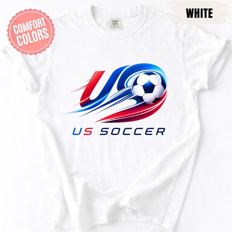 K&ouml;nnte beinhalten: Ein wei&szlig;es Kurzarm-T-Shirt mit einer Grafik eines Fu&szlig;balls mit roten und blauen Bewegungsstreifen. Das Design enth&auml;lt den Text US SOCCER in blauer und roter Schrift. Ein rosa kreisf&ouml;rmiges Abzeichen zeigt COMFORT COLORS und WHITE.