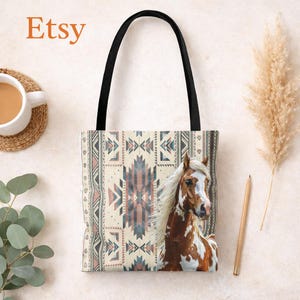 Peut inclure: Sac fourre-tout avec une anse noire et un fond crème. Le sac présente un motif géométrique d'inspiration amérindienne dans des tons de bleu, rose et marron. Un cheval brun et blanc avec une crinière blanche flottante est sur le côté droit. Le mot "Etsy" est dans le coin supérieur gauche.