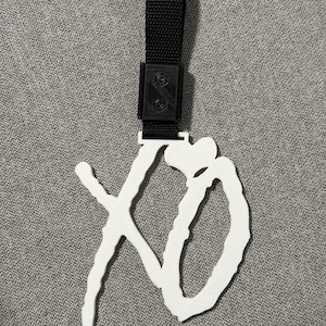 May include: White 3D-printed XO logo keychain with a black strap. The keychain features a stylized "XO" design, with the letters connected and a heart shape incorporated. The black strap is attached to the top of the logo.