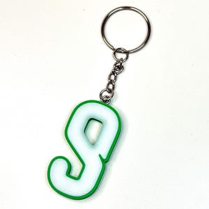 May include: A keychain featuring a white number nine with a vibrant green border. The number is attached to a silver-colored metal split ring by a short metal chain. The design has a clean, bold aesthetic with a smooth finish on the white surface.