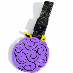 Puede incluir: Un colgante circular de color morado con un patrón de remolinos en relieve en toda su superficie. Una pequeña pieza amarilla se sitúa en la parte superior, conectada a un enganche de plástico negro y a una correa de tela negra para colgar.