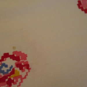 Pu&ograve; includere: Primo piano di un tessuto color crema con motivi floreali ricamati a punto croce in tonalit&agrave; rosa, rosso, verde, giallo e blu. Il design &egrave; pixelato, conferendo un'estetica vintage o fatta a mano. Il tessuto sembra essere cotone o lino.