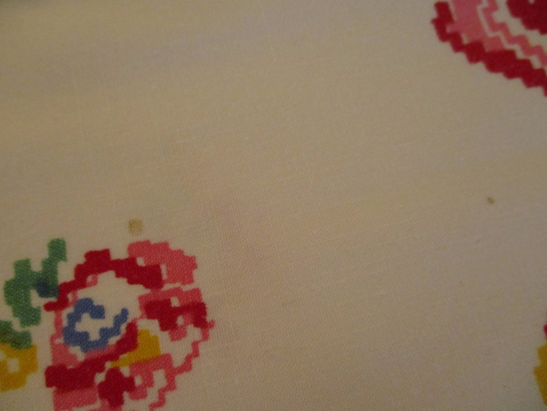 Pu&ograve; includere: Primo piano di un tessuto color crema con motivi floreali ricamati a punto croce in tonalit&agrave; rosa, rosso, verde, giallo e blu. Il design &egrave; pixelato, conferendo un'estetica vintage o fatta a mano. Il tessuto sembra essere cotone o lino.