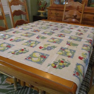 May include: A rectangular tablecloth featuring a repeating grid pattern of square blocks on a light beige background. Each block contains illustrations of yellow lemons, green leaves, and small red flowers framed by a blue border with white accents.
