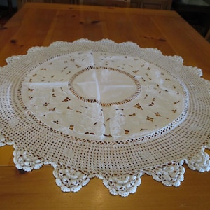 May include: A round white doily featuring a central fabric panel with cutwork embroidery, surrounded by a wide crocheted border with a scalloped edge. The intricate needlework displays floral motifs and geometric patterns in white thread.