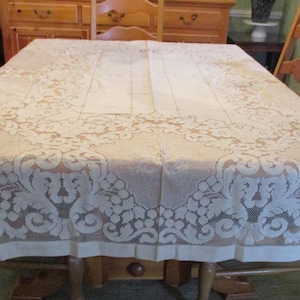 May include: A beige rectangular tablecloth with intricate floral lace detailing. The tablecloth is draped over a wooden table, with wooden chairs visible on either side. The lace design is prominent along the edges.