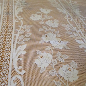 May include: Cream-colored lace fabric featuring a floral rose pattern. The lace has a delicate, open weave with detailed rose and leaf designs. Vertical borders with geometric and floral patterns frame the floral design.