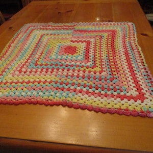 May include: A handmade, square crochet blanket in a granny stitch pattern. The blanket features a vibrant color palette of pink, yellow, blue, and white. The blanket is approximately 40 inches square, with a scalloped edge.
